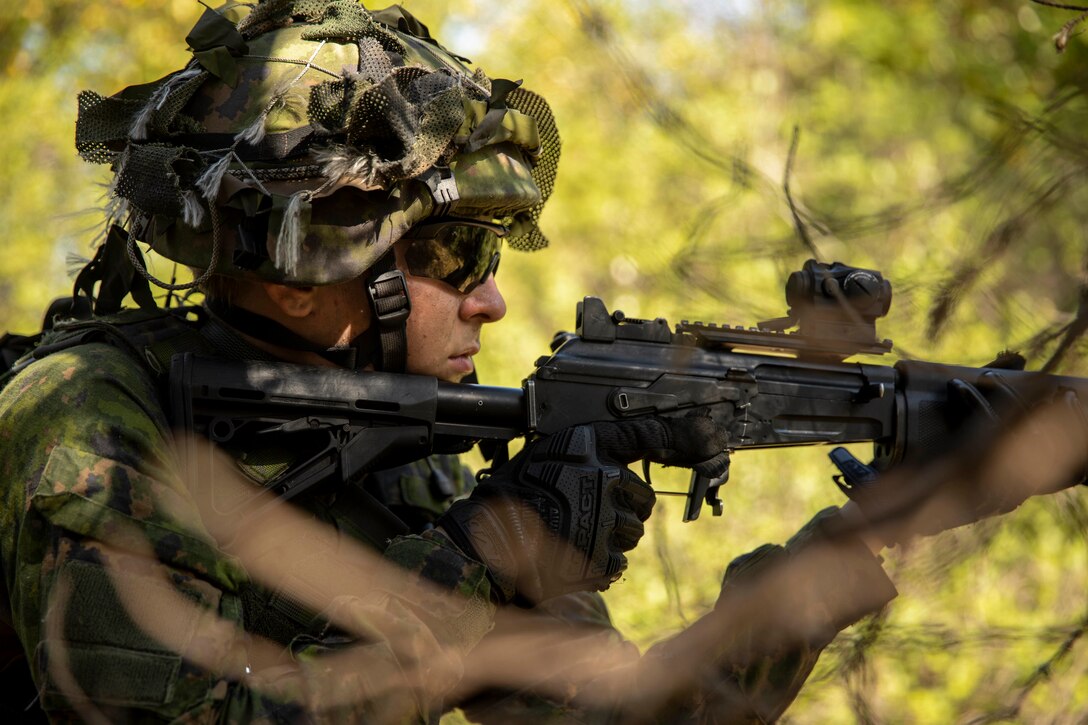 A soldier aims his weapon in a wooded setting.