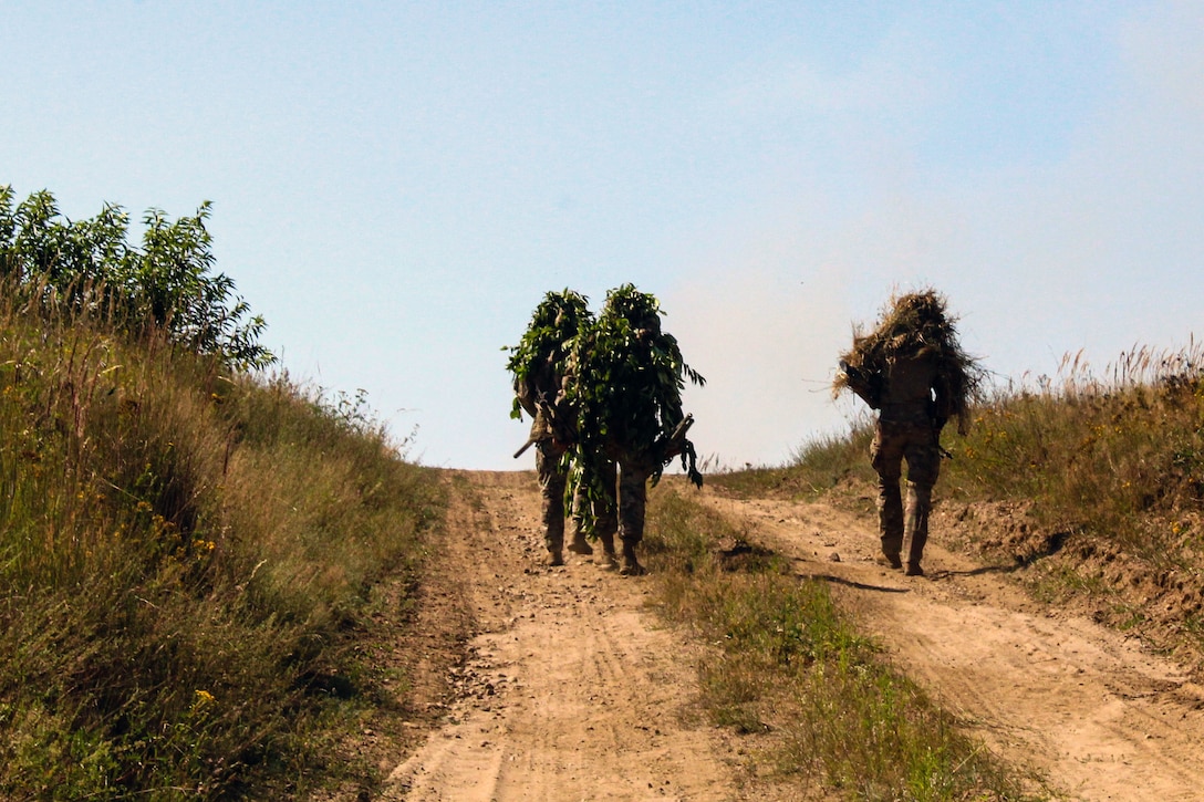 Three soldiers wearing backs covered in grass carry weapons and walk up a hill.