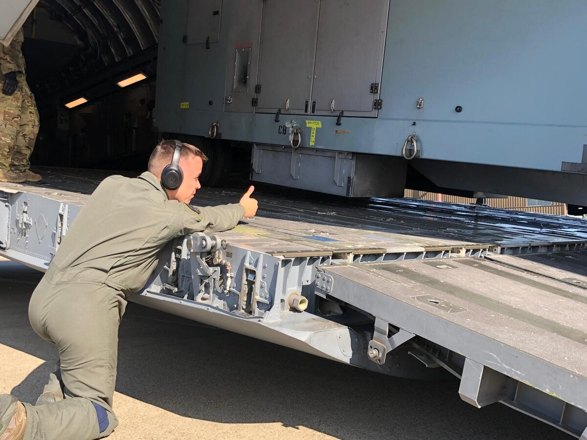 man in flight suit on one knee giving thumbs up as large generator is loaded onto aircraft ramp