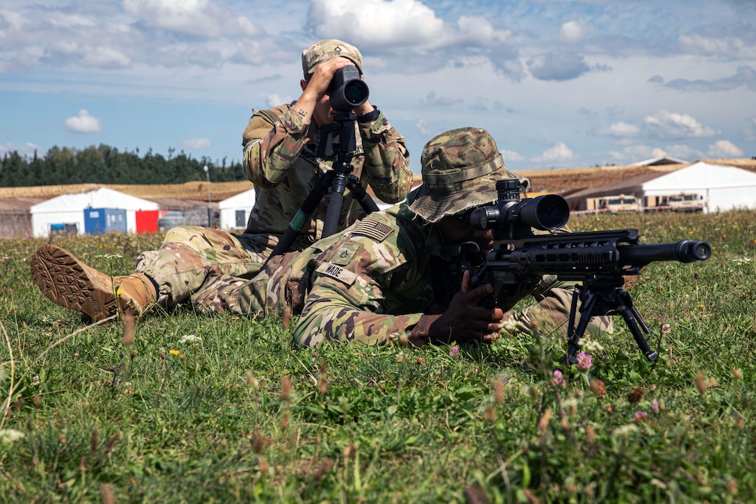 One soldier looking through binoculars and another soldier looks through the scope of a weapon while conducting sniper training.