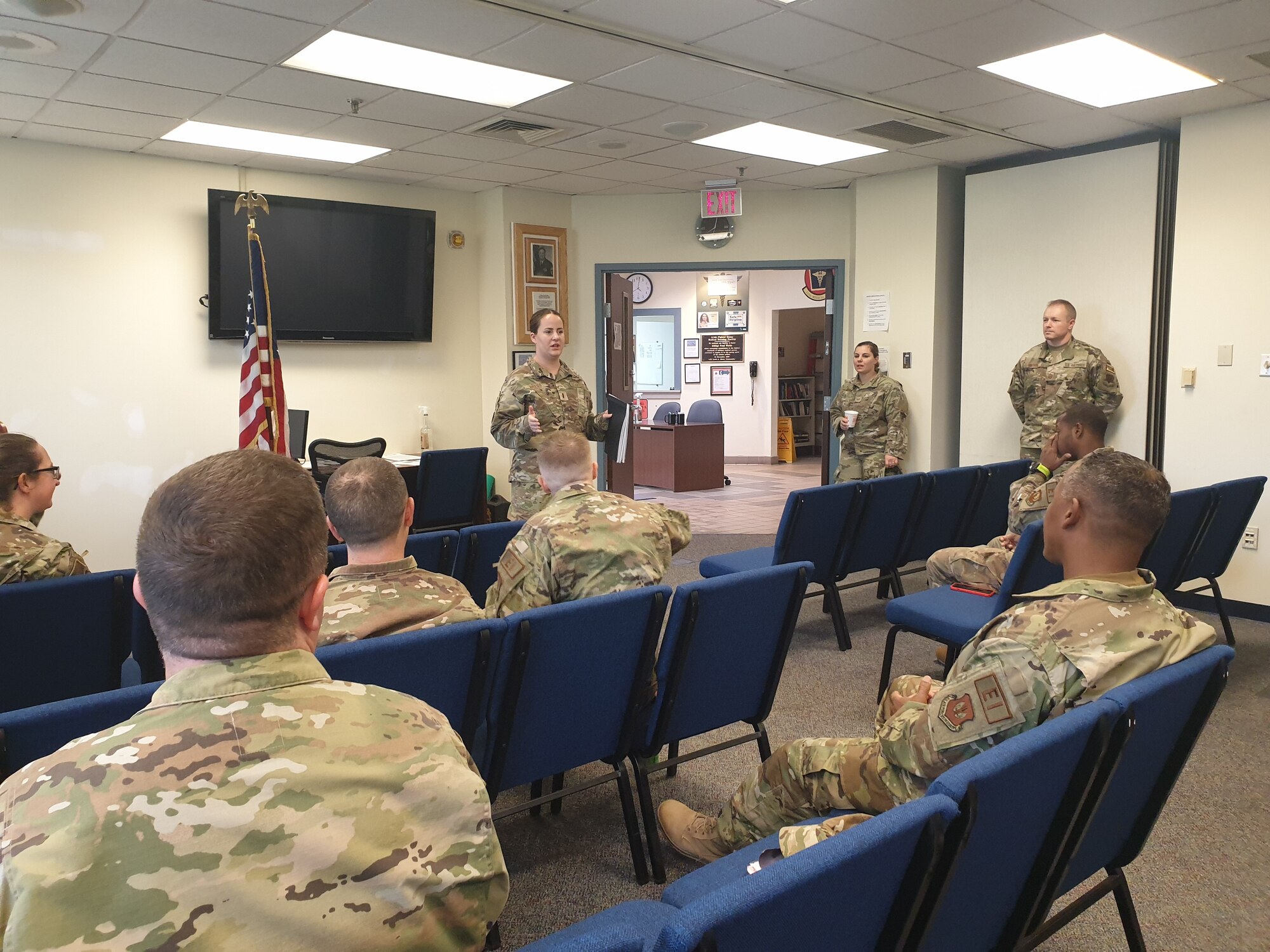 Woman in military uniform talks to seated military personnel.