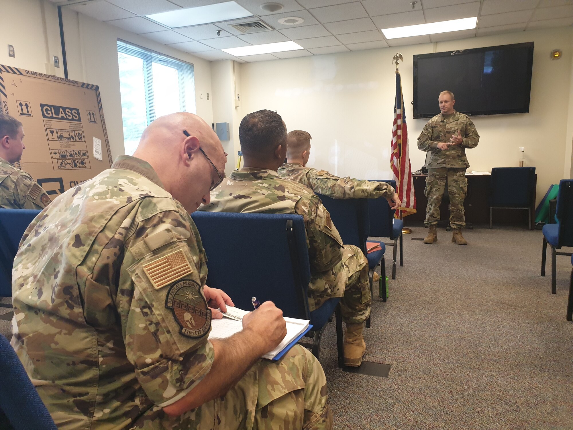 Man in military uniform talking to seated military personnel.