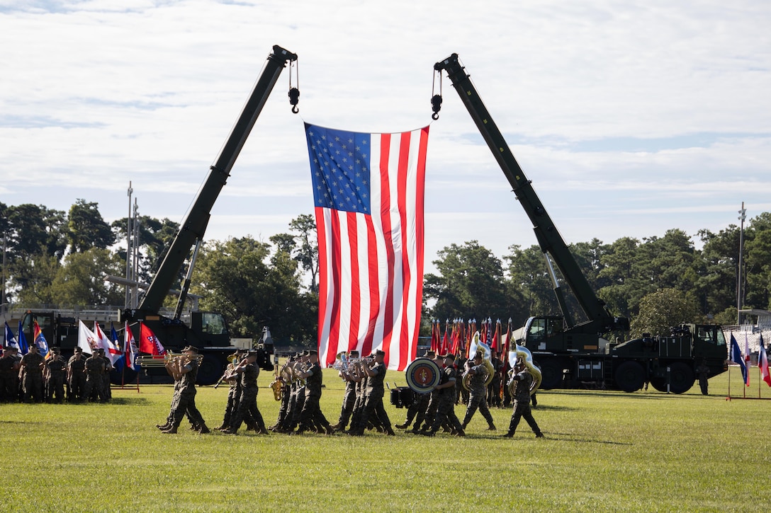 Band members perform next to a large American flag displayed by two cranes on a field where others stand holding flags.