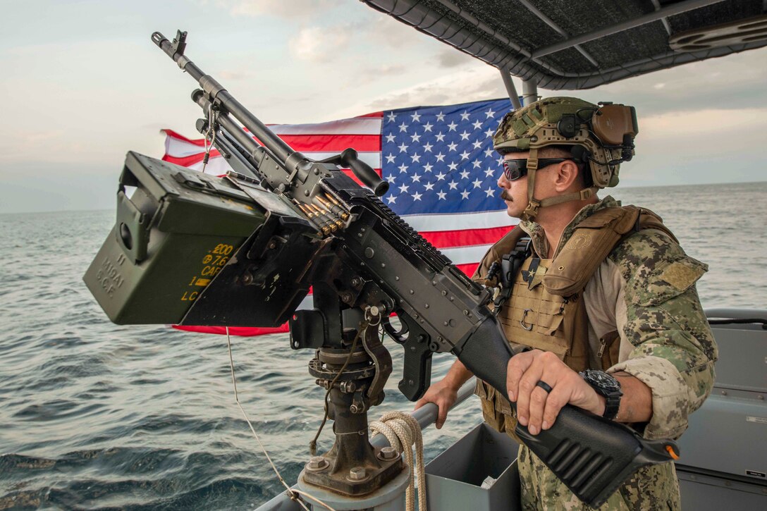 A sailor holds a weapon while standing in front of an American flag on a boat at sea.