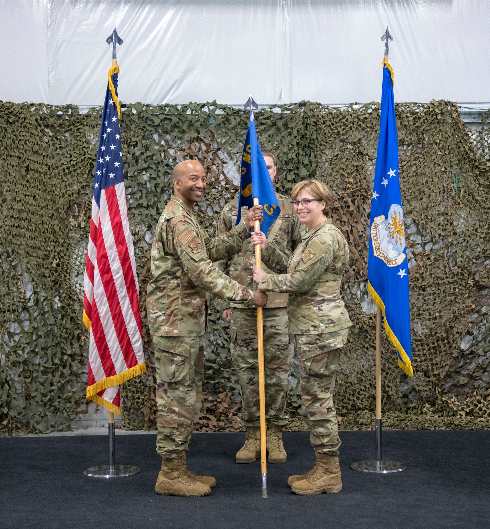 Col. Dorroh passes the 446th Civil Engineer Squadron guidon to Lt. Col. Hartman in operational camouflaged pattern uniforms in front of camo netting backdrop.
