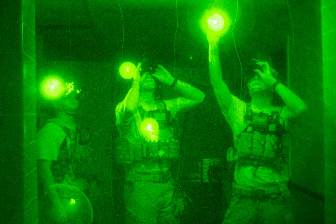 Three airmen illuminated by green light look up while wearing goggles.