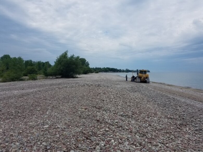 Wide photo of a stone beach with trees on the left, water on the right and a person standing next to a forklift discussing matters with the forklift driver.