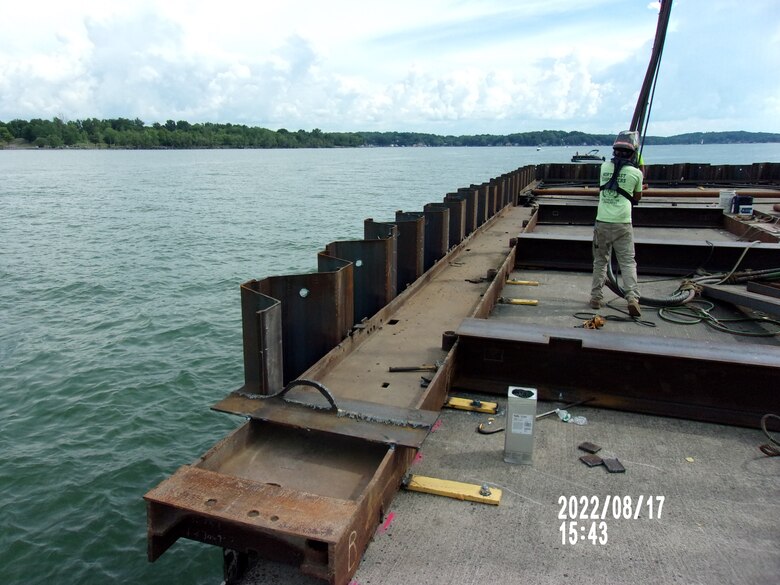 A worker stands on the pier amongst steel beams as the water of the bay surrounds him, holding the coupling needed to perform work of the day.