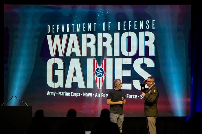 A general salutes Jon Stewart on a stage with the Warrior Games logo on a large monitor.
