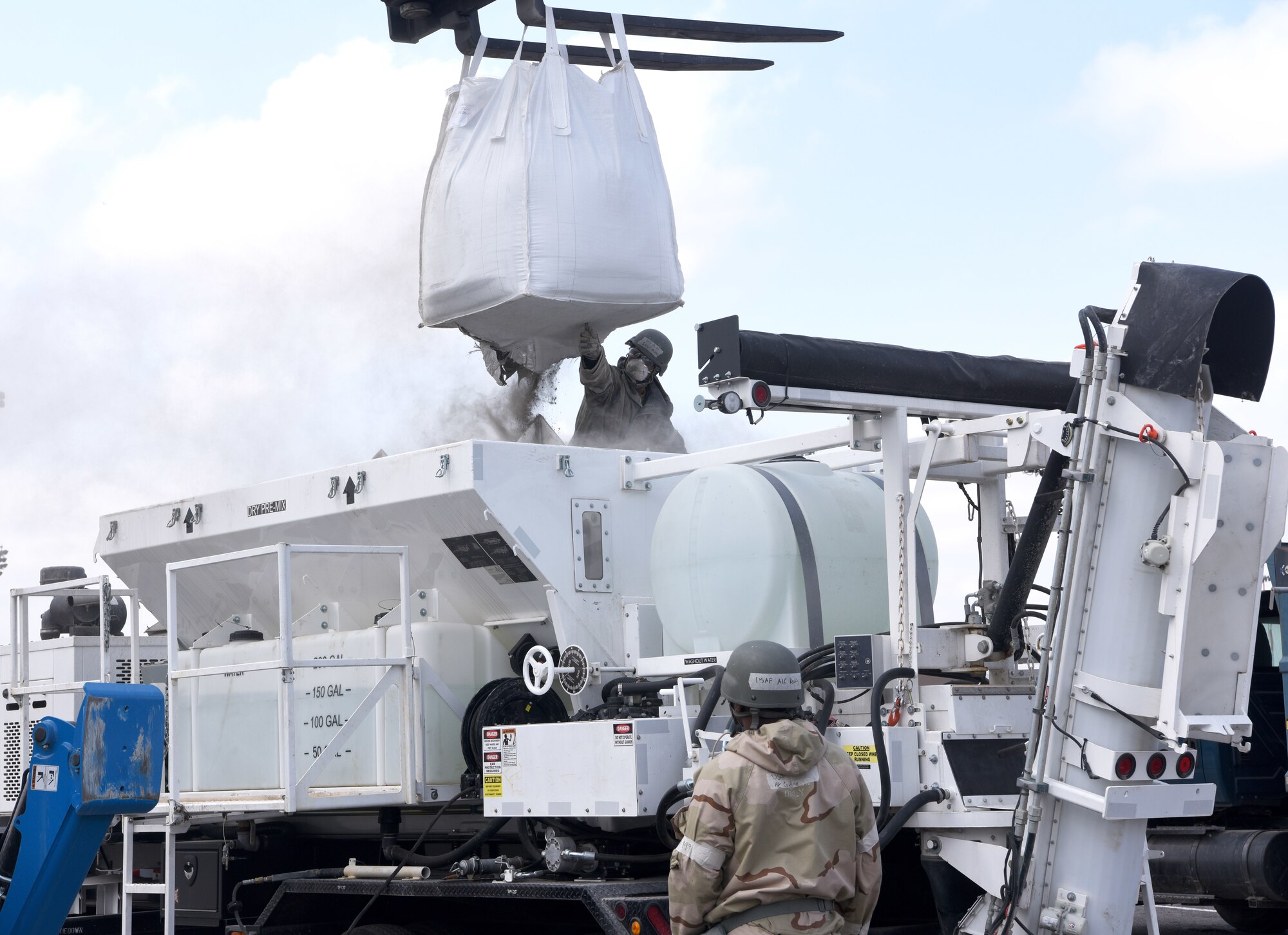 U.S. Air Force Airman 1st Class Jahsiah Keating, on top of vehicle, cuts open a bag of cement ready for a procedure known as “slash and splash” – slash open a bag of cement and splash with water – ready to pour it into a large, pre-cut hole in a simulated runway during a simulated damaged runway repair as part of a chemical, biological, radiological, nuclear and explosives exercise at Royal Air Force Mildenhall, England, Aug. 17, 2022. The 100th CES Airmen used a variety of equipment including excavators and loaders to practice removing damaged concrete and base course on a simulated runway. They practiced repair, break up and removal of concrete, before mixing and pouring new, quick-drying concrete and levelling it. (U.S. Air Force photo by Karen Abeyasekere)