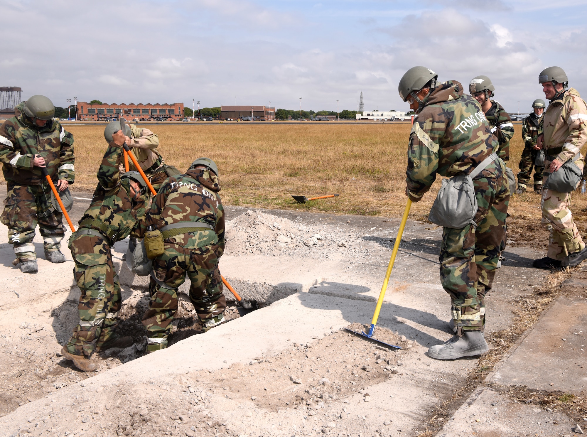 U.S. Air Force Airmen from the 100th Civil Engineer Squadron clear broken concrete during a simulated damaged runway repair as part of a chemical, biological, radiological, nuclear and explosives exercise at Royal Air Force Mildenhall, England, Aug. 17, 2022. The 100th CES Airmen used a variety of equipment including excavators and loaders to practice removing damaged concrete and base course on a simulated runway. The 100th Air Refueling Wing commander, Col. Gene Jacobus, right, visited the Airmen to watch as they practiced repair, break up and removal of concrete, before mixing and pouring new, quick-drying concrete and levelling it. (U.S. Air Force photo by Karen Abeyasekere)