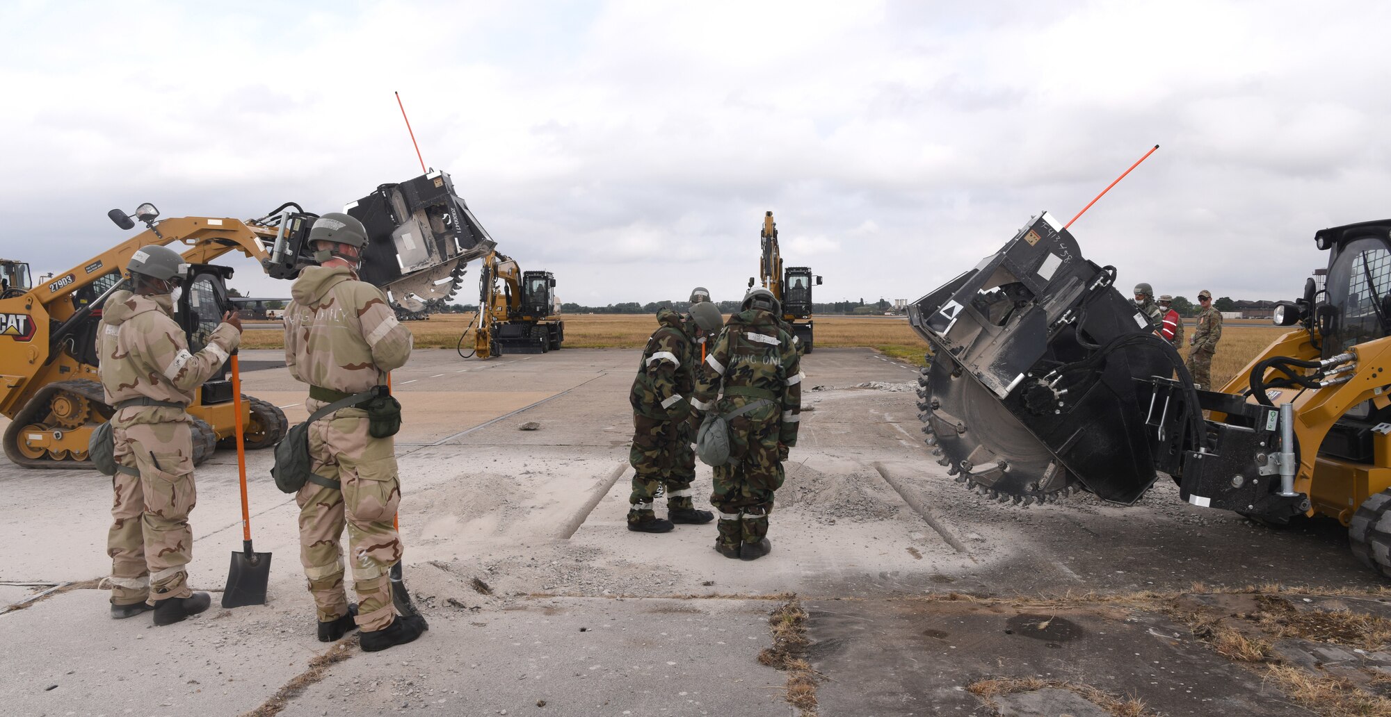 U.S. Air Force Airmen from the 100th Civil Engineer Squadron prepare to clear broken concrete cut by two compact track loaders during a simulated runway repair as part of a a chemical, biological, radiological, nuclear and explosives exercise at Royal Air Force Mildenhall, England, Aug. 17, 2022. The Airmen used a variety of equipment including excavators and loaders to practice removing damaged concrete and base course on a simulated runway. They practiced repair, break up and removal of concrete, before mixing and pouring new, quick-drying concrete and levelling it, all while wearing mission-oriented protective postures gear. (U.S. Air Force photo by Karen Abeyasekere)