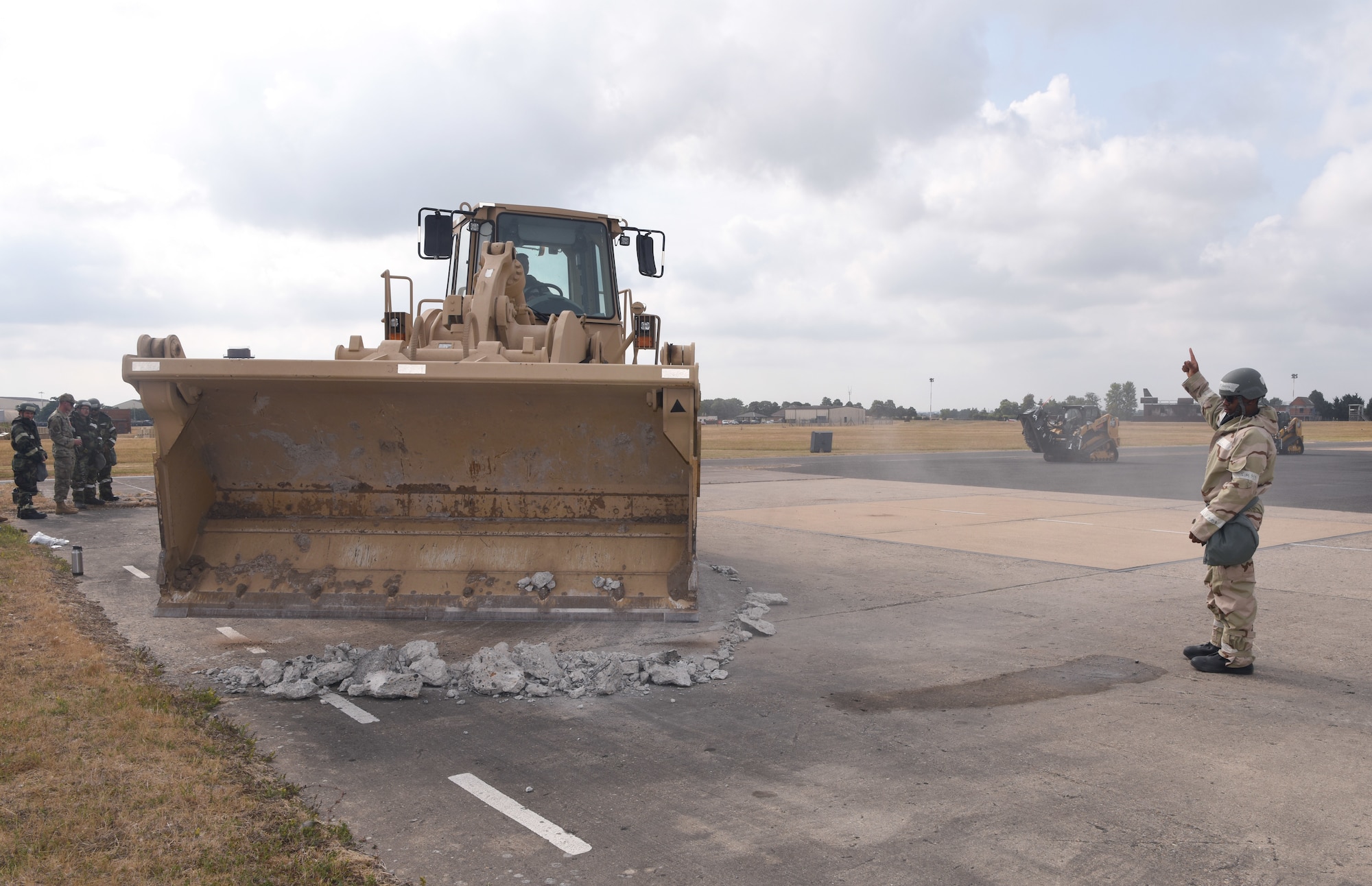 U.S. Air Force Airman 1st Class Jahsiah Keating, right, 100th Civil Engineer Squadron pavement and equipment journeyman, directs a loader driven by Tech. Sgt. John Barber, 100th CES Power Production NCO in charge, to clear lumps of broken concrete during a chemical, biological, radiological, nuclear and explosives exercise at Royal Air Force Mildenhall, England, Aug. 17, 2022. The Airmen used a variety of equipment including excavators and loaders to practice removing damaged concrete and base course on a simulated runway. They practiced repair, break up and removal of concrete, before mixing and pouring new, quick-drying concrete and levelling it, all while wearing mission-oriented protective postures gear. (U.S. Air Force photo by Karen Abeyasekere)