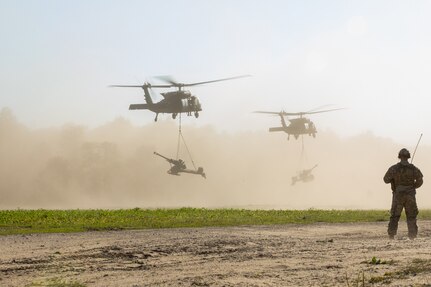 An Ohio Army National Guard Soldier watches UH-60 Black Hawks, assigned to 1st General Support Aviation Battalion, 111th Aviation Regiment, carry M119A3 Howitzers, belonging to 1st Battalion, 134th Field Artillery Regiment, during Operation Northern Strike at Camp Grayling, Mich., Aug. 8, 2022.