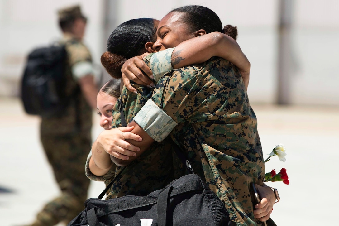 Three Marines hug one another while carrying a shoulder bag and roses.