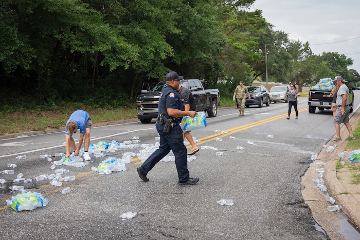 A small crowd picks fallen water bottles off of a suburban street while a police officer walks across the scene carrying a case of the fallen bottles.