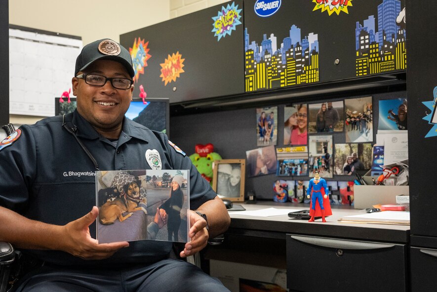 Police officer sits at his desk. The desk is covered with brightly covered comic art and a superman figurine is posed on the desk. The police officer is holding a photo of his wife and two dogs.