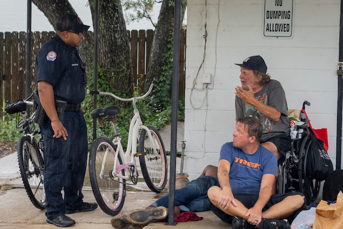 Police officer and two homeless men speak under building overhang.