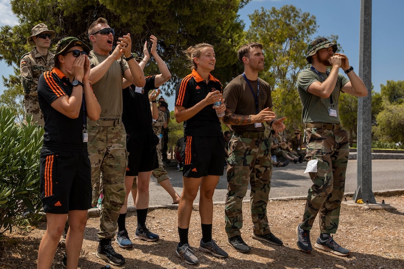 Service members stand in a row outdoors and cheer, some in athletic garb.