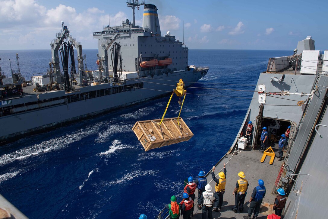 A large box of supplies is cabled over water between two ships.
