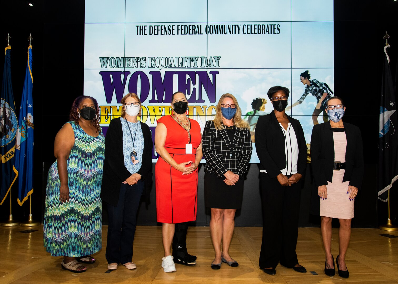 Six women stand on stage with masks on. A heavyset  African American woman in a patterned dress, a woman with red hair and glasses wearing a black suit and blue blouse, a African American woman with black hair pulled back in a red dress in a walking boot, a woman with blond hair and glasses wearing a checkered suit jacket and black skirt, a African American woman with short black hair in a black suit with a white blouse and a woman in glasses and dark hair pulled back in a bun wearing a pink and black dress with a black jacket.