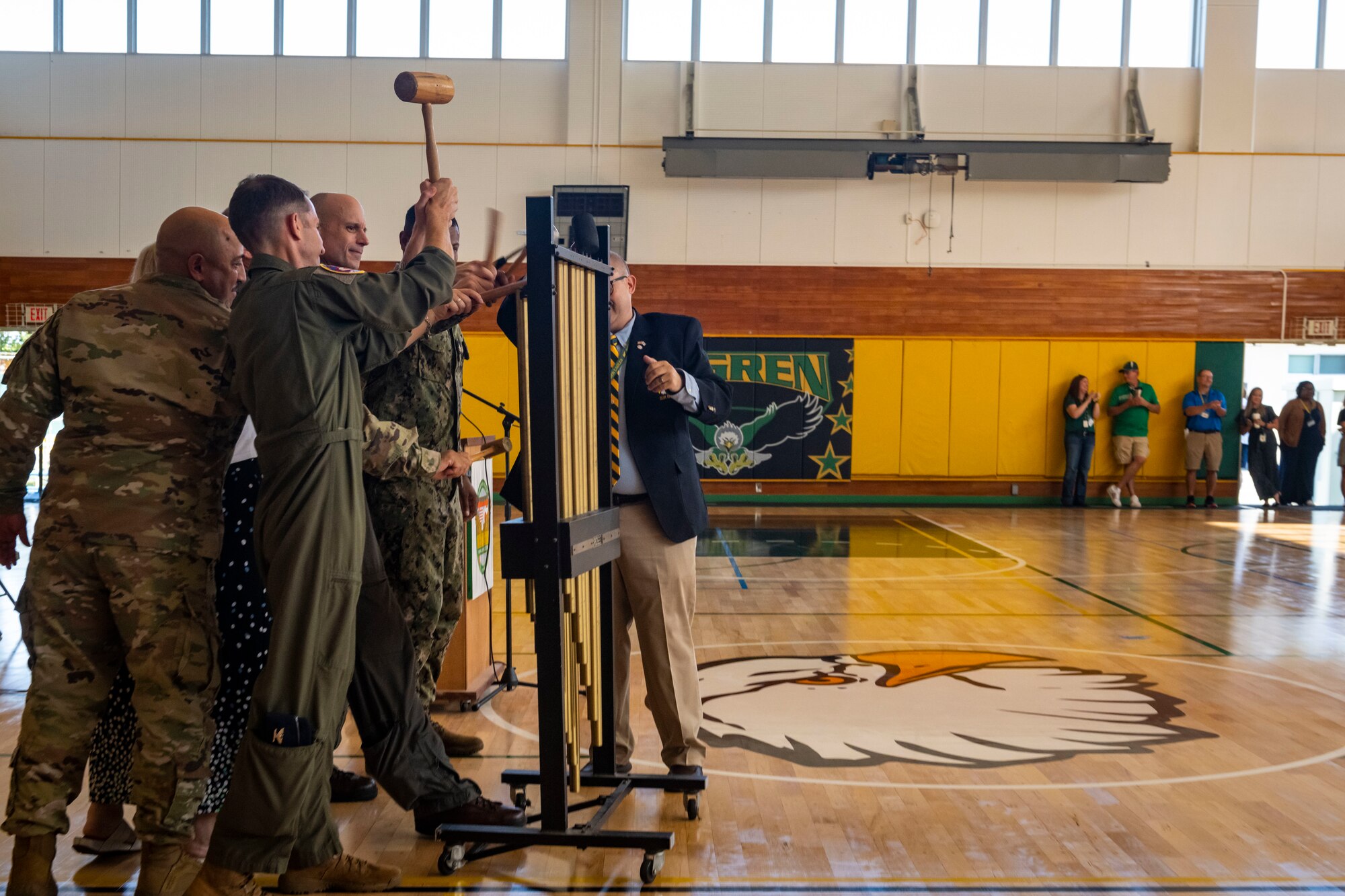 Military members in uniform swing mallets at the tubular bell instrument in a gym