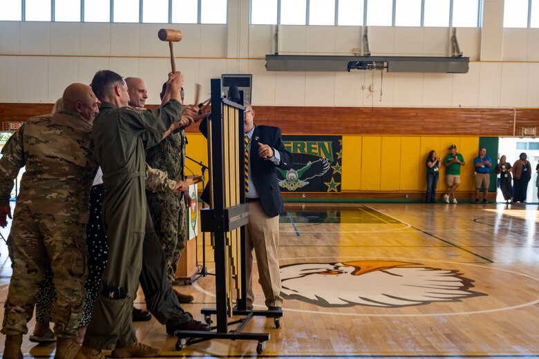 Military members in uniform swing mallets at the tubular bell instrument in a gym