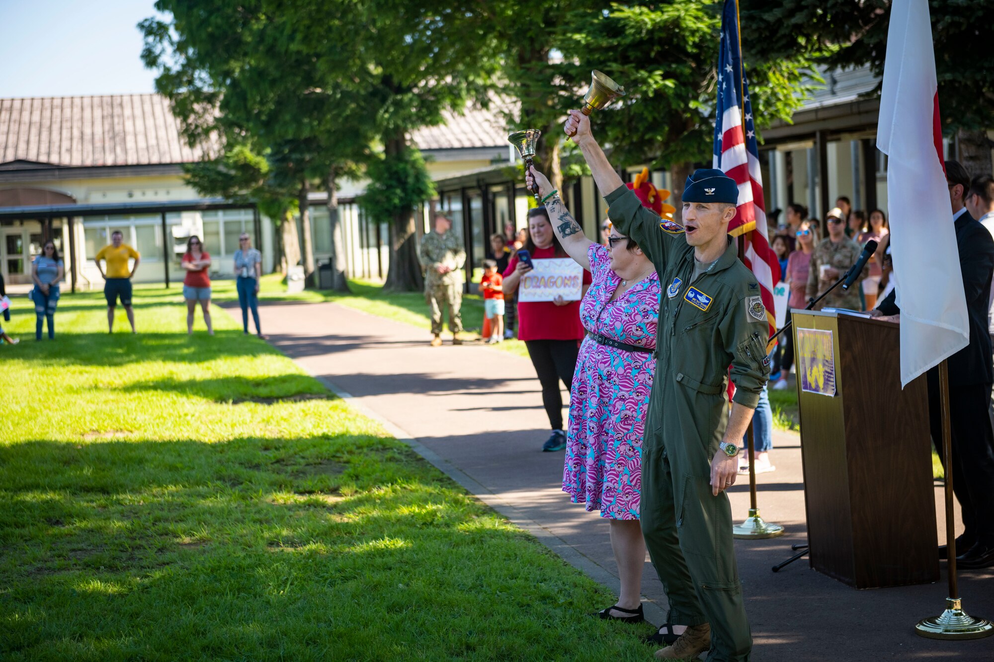 Military member in uniform and civilian both shake bells above their heads in front of a crowd