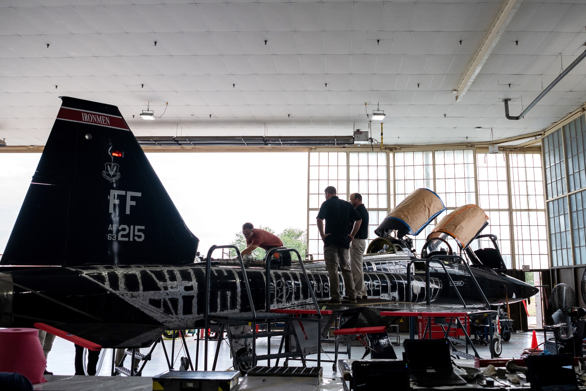 Three men standing on fighter jet in maintenance hangar