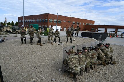 Soldiers in a rank crouch behind their shields as a group of other soldiers stand in front of them and throw stones