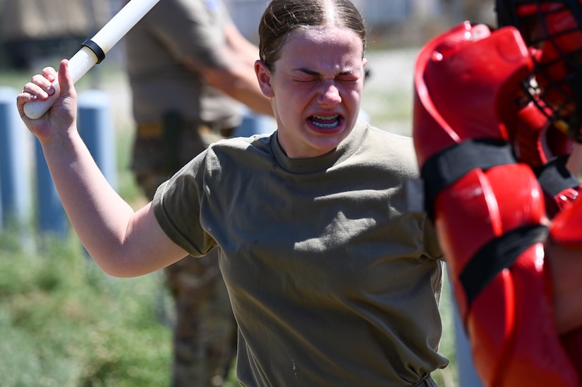A female soldier with her eyes nearly closed due to being sprayed with OC spray, uses a baton to engage a simulated adversary .