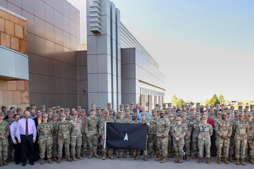 A group of service members and civilians pose next to building.