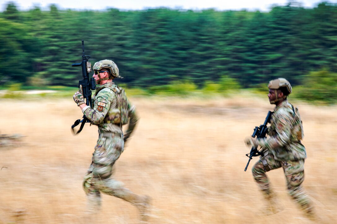 Two airmen run through field of tall grass while carrying weapons.