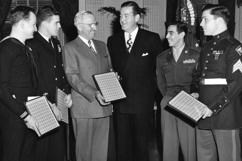 Six men stand together, with four holding sheets of stamps.