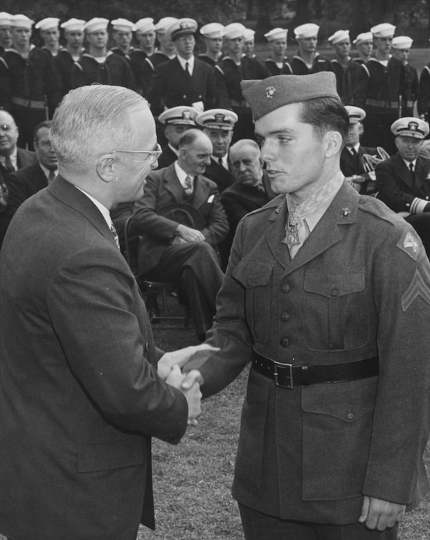 Two men shake hands while many others sit in the background.