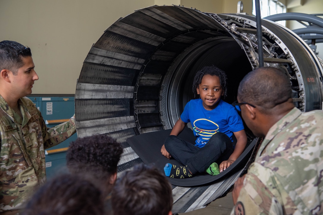 A child sits inside a jet engine as service members gather around.