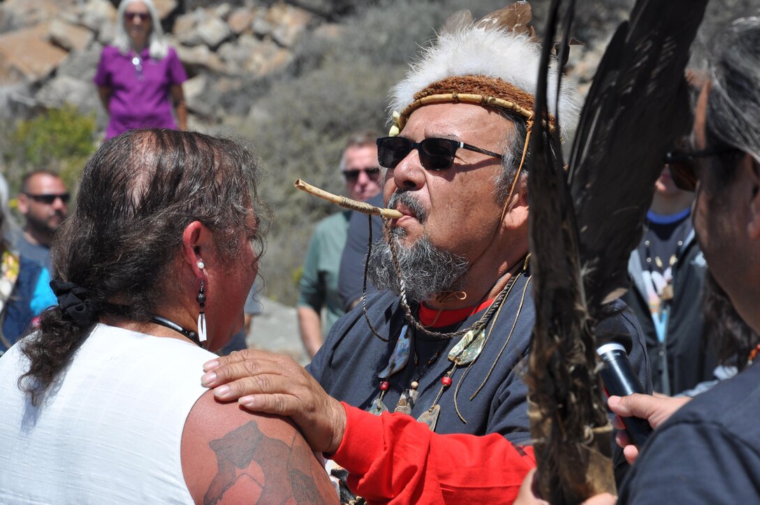 A Chumash tribal elder blows a reed whistle as he blesses a woman circled by other tribe members. He wears sunglasses and tribal headgear, his tilted skyward as he places his hands on her shoulders.