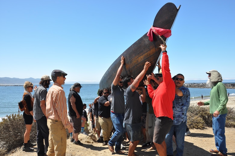 A long, heavy, seagoing boat called a tomol is lifted by volunteers from the rocky shoreline to the base of Morro Rock. The weight of the dark planked boat is evident in the faces of the volunteers as it crests the shoreline boulders. A