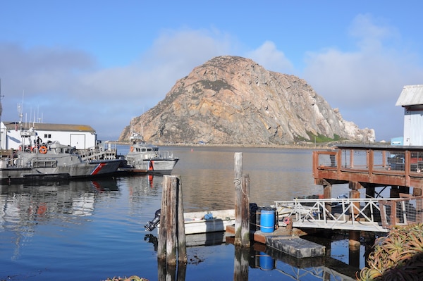 A giant rock once a volcanic core towers over Morro Bay. The Chumash Indian event, Reunite the Rock, can be seen as tiny figures at its base. A thick wall of fog looms a few miles off the coast. Morro Bay itself is bathed in sunlight.