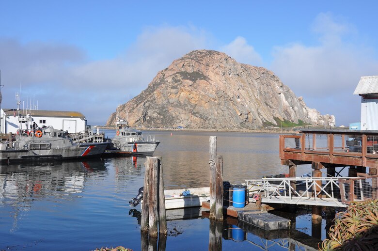 A giant rock once a volcanic core towers over Morro Bay. The Chumash Indian event, Reunite the Rock, can be seen as tiny figures at its base. A thick wall of fog looms a few miles off the coast. Morro Bay itself is bathed in sunlight.