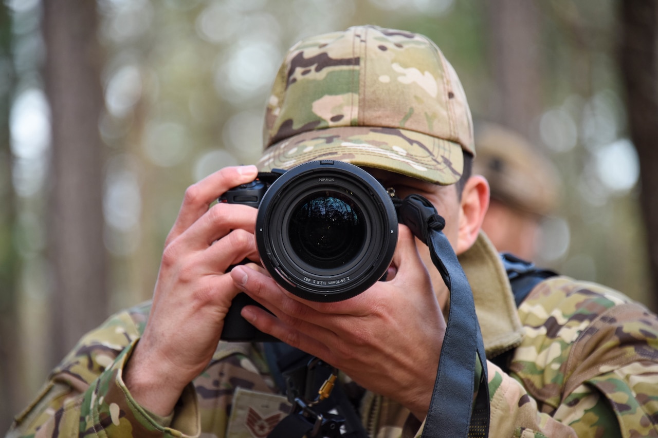 An airman looks through a camera lens in a forested area.