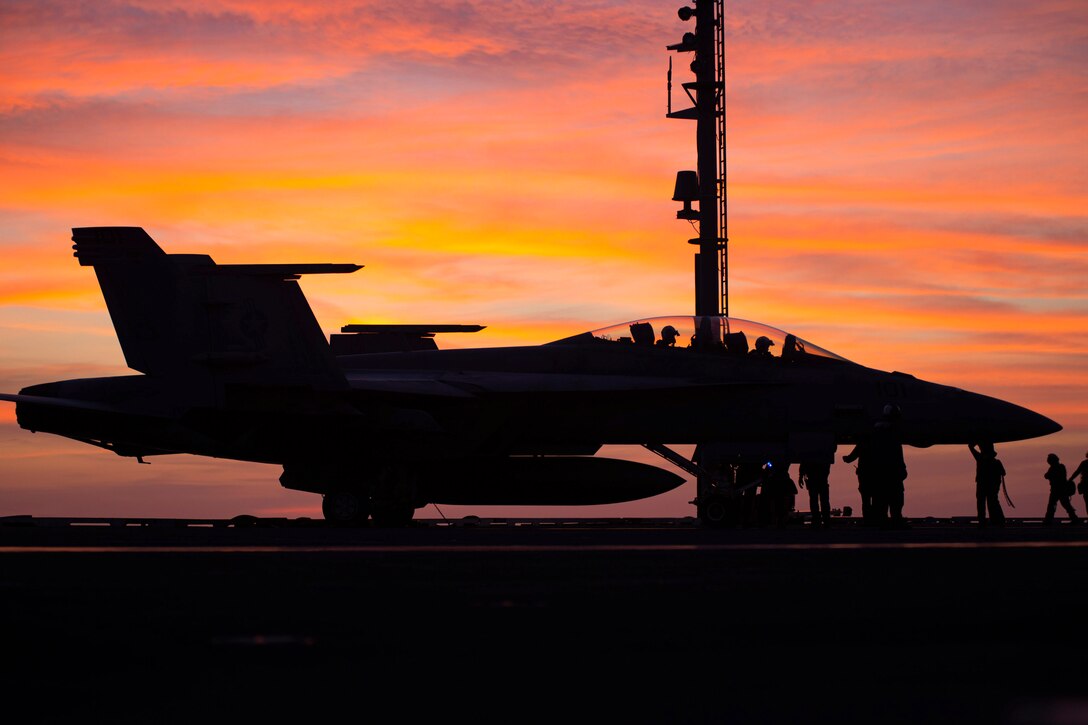 An aircraft is parked on a tarmac aboard a ship with sailors standing nearby under a sunlit sky.