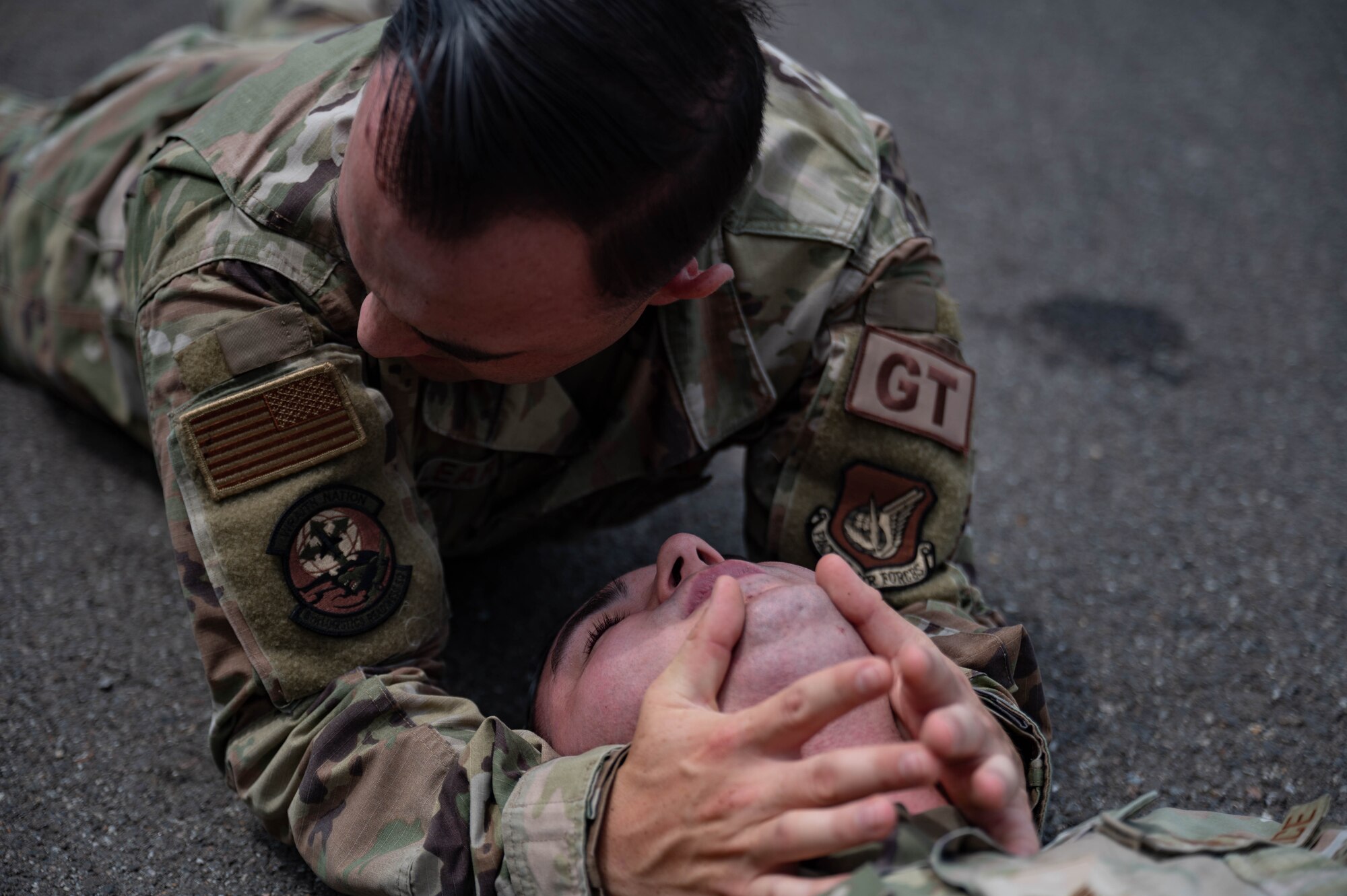Airman lies on a ground during simulated medical event while another Airman assists by providing aide.