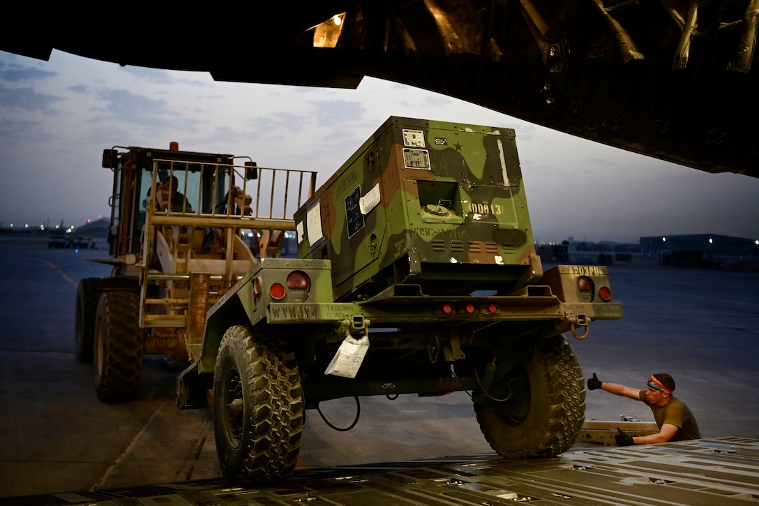 An airman standing at the foot of a ramp gives hand directions to an airman driving a forklift loaded with cargo.