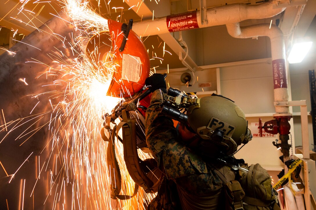 Sparks fly as a Marine wearing protective gear uses a saw to open a metal door.