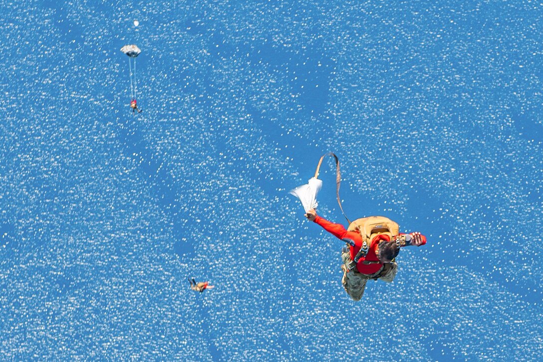 An airman releases his parachute as fellow airmen free fall over a body of water in the background.