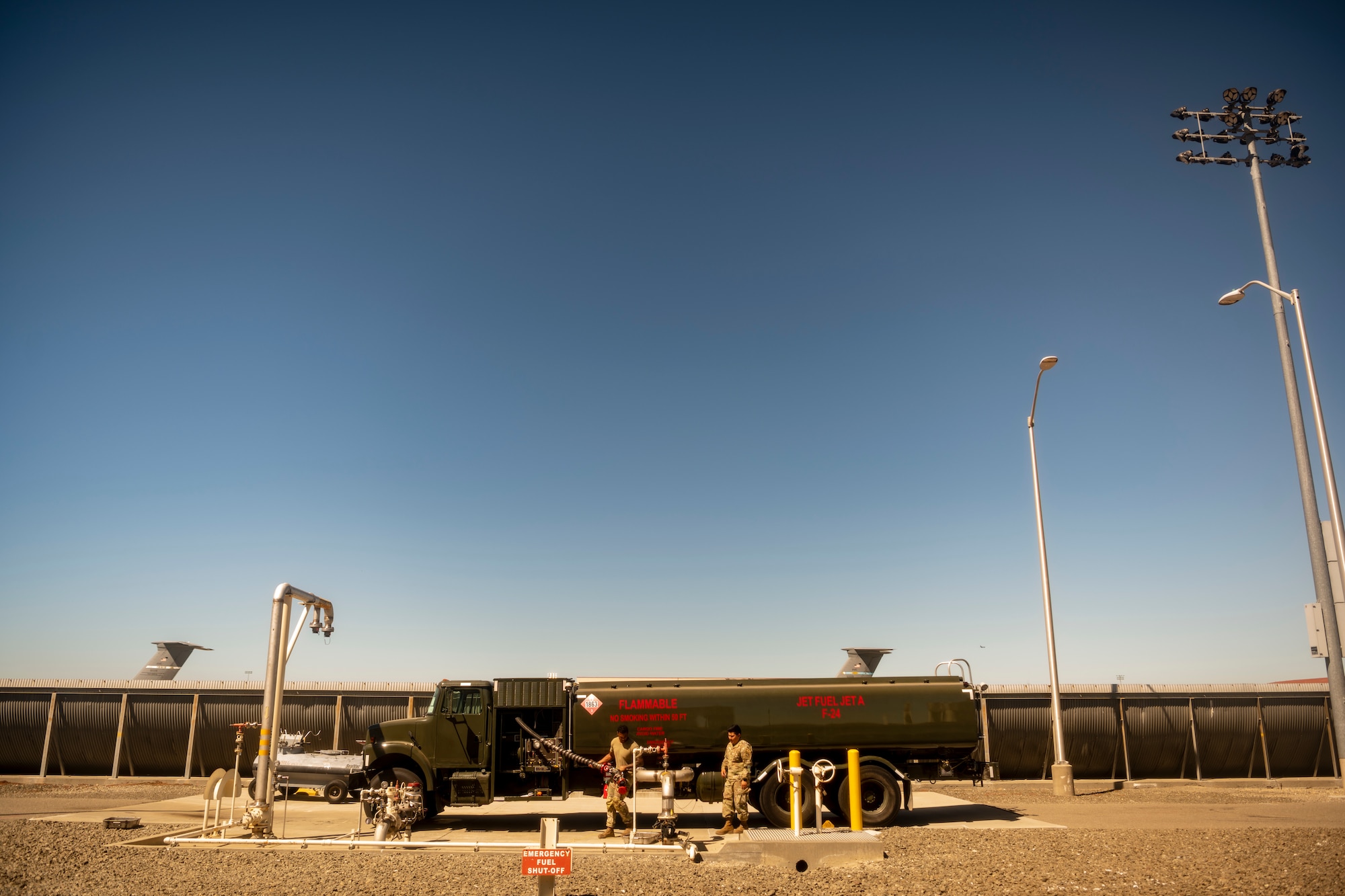 a green truck with a massive tank on the back, and two men in front of the truck move equipment on the truck to prepare the truck to transfer all of its fuel.