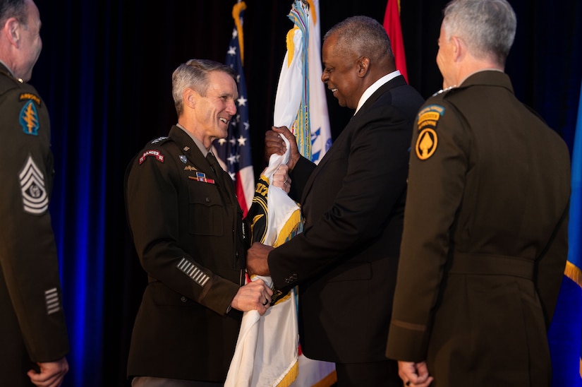 Secretary of Defense Lloyd J. Austin III hands a flag to a general while two others look on.