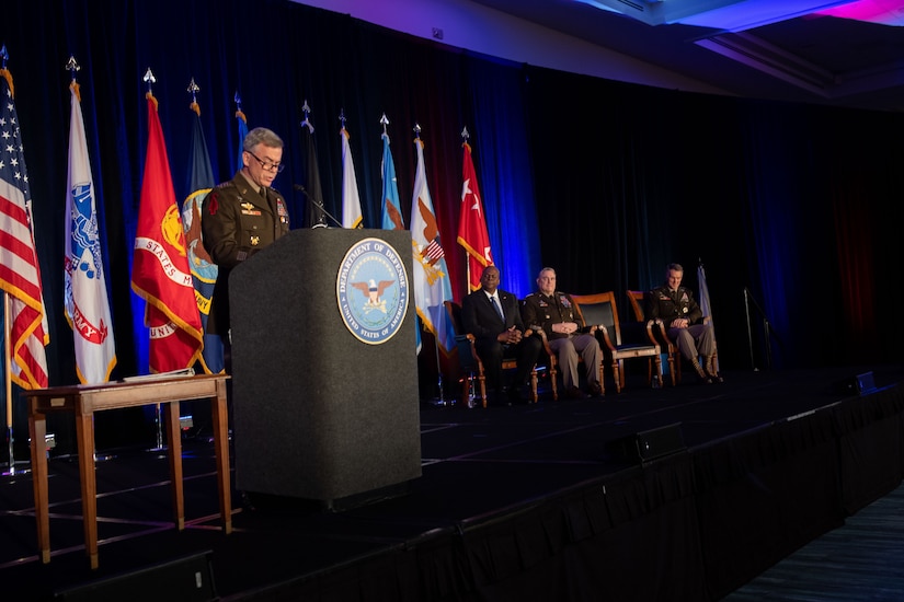 A general speaks at a podium while three other people on stage look on.