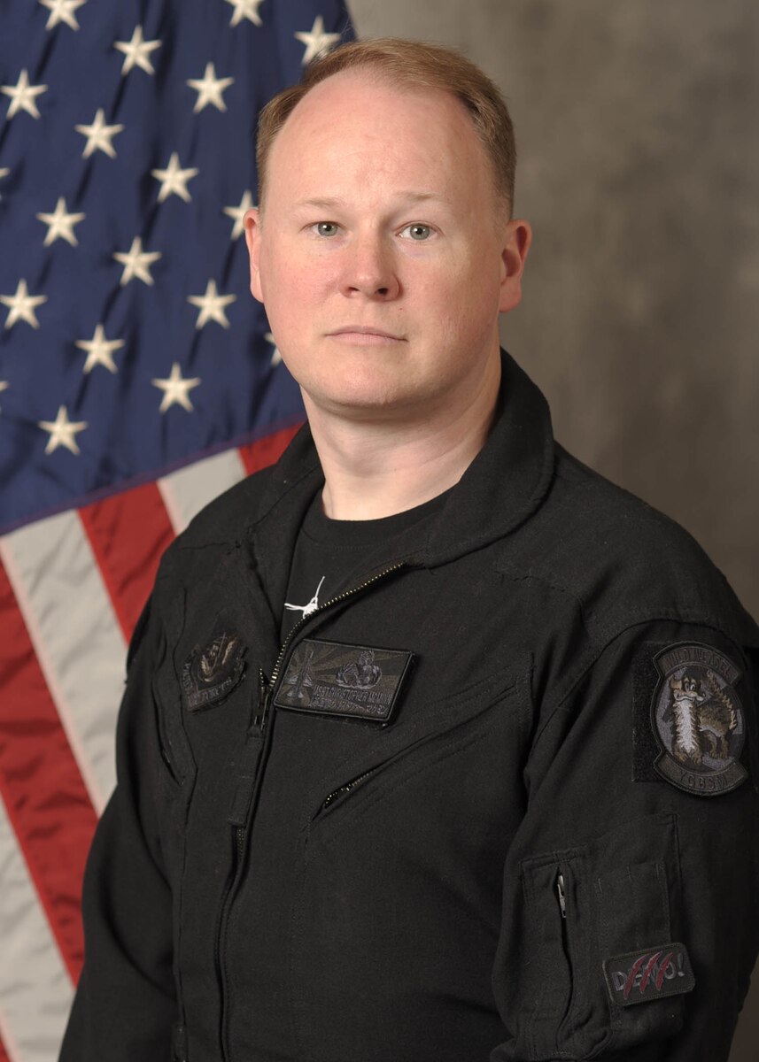 A man in a black flight suit in front of an American Flag